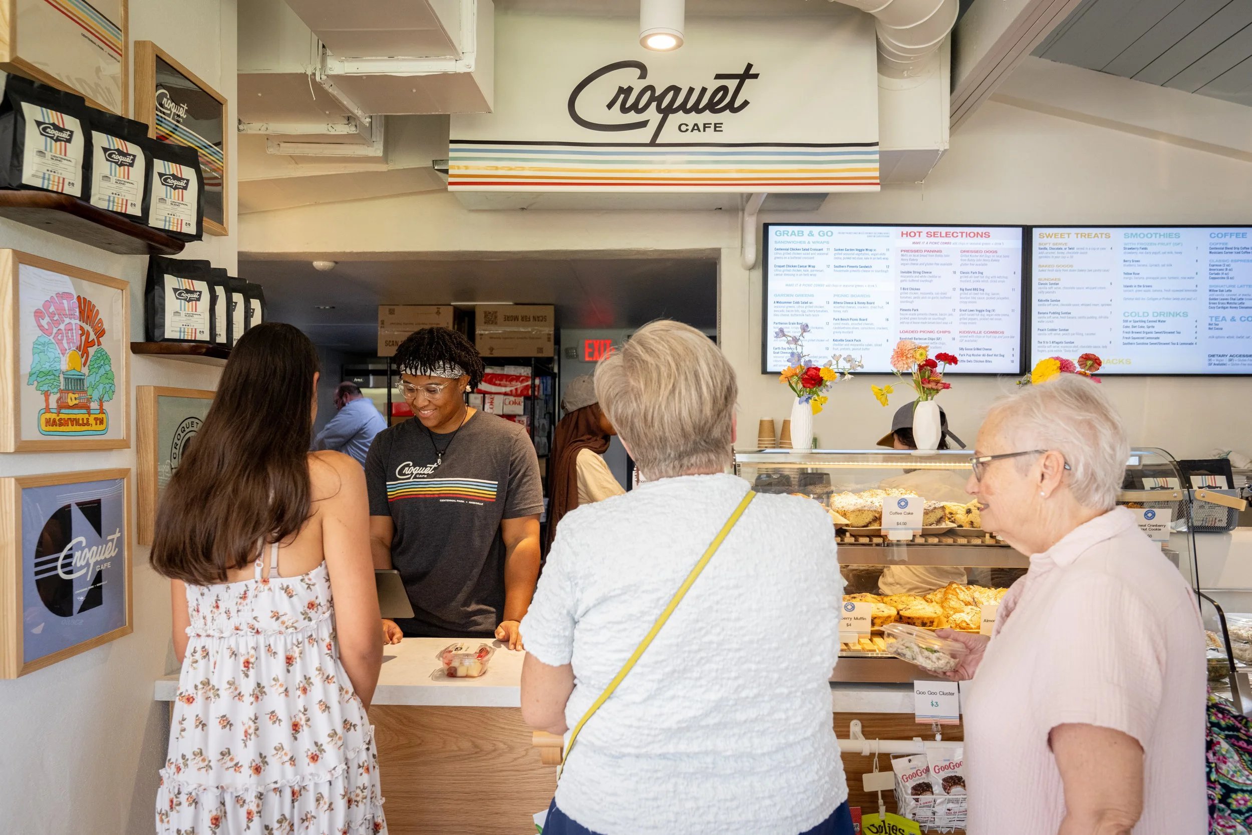 Inside Croquet Cafe on grand opening day: customers at the counter under the cafe's sign, with menu boards and a pastry case