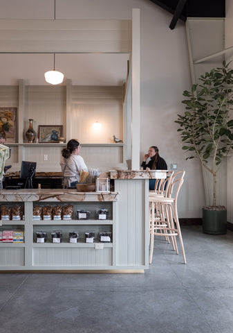 Inside a Perenn market: a marble-topped counter with packaged goods shelved below, staff at work behind it, and café seating beside a tall potted tree